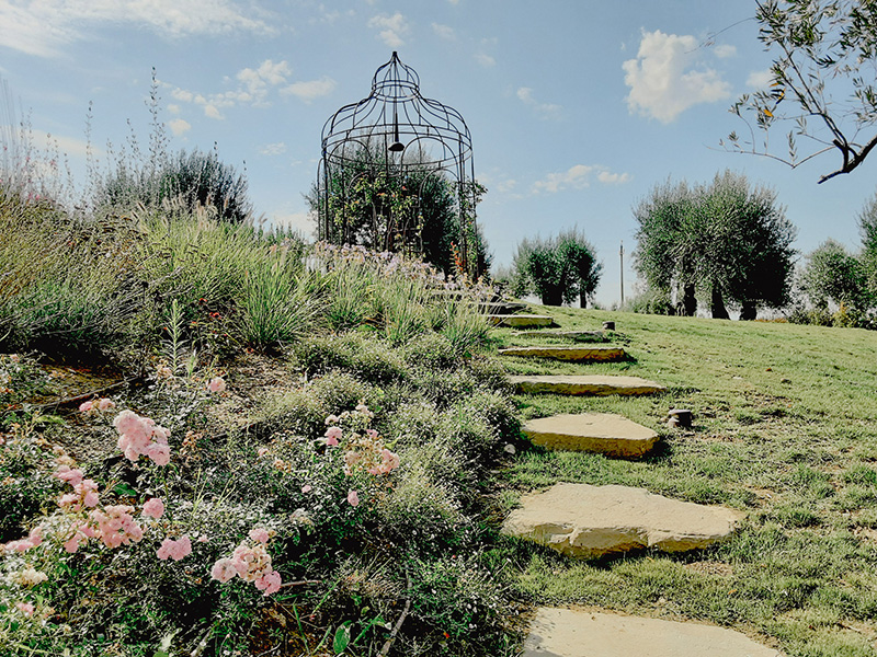 Giardino con fiori, un sentiero di rocce in salita su un grande prato verde e un gazebo in ferro battuto in lontananza.